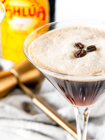 Espresso martini cocktail with foam and three coffee beans in glass close up with kahlua bottle in the background