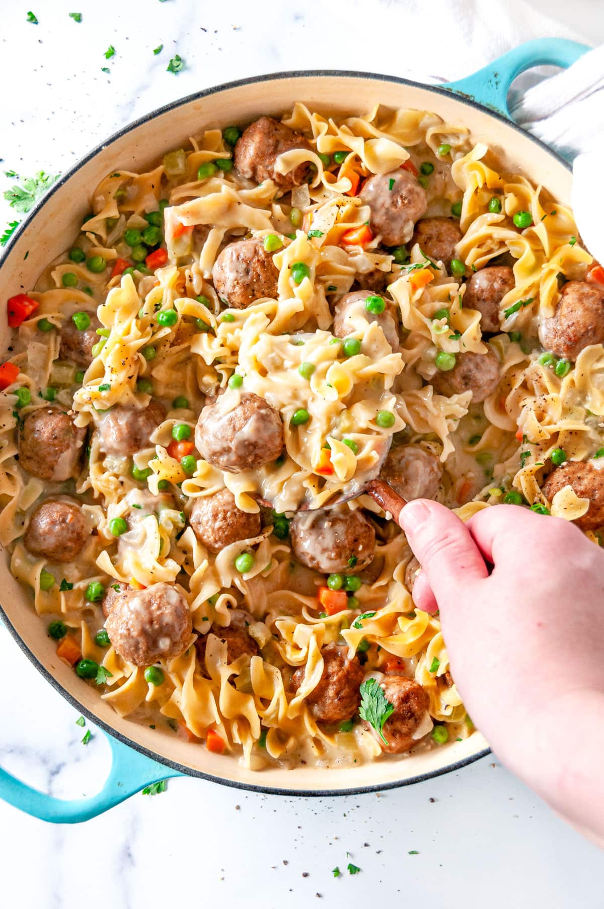 One Pot Swedish Meatball Pasta in blue le creuset braiser with wooden spoon held in hand on white marble