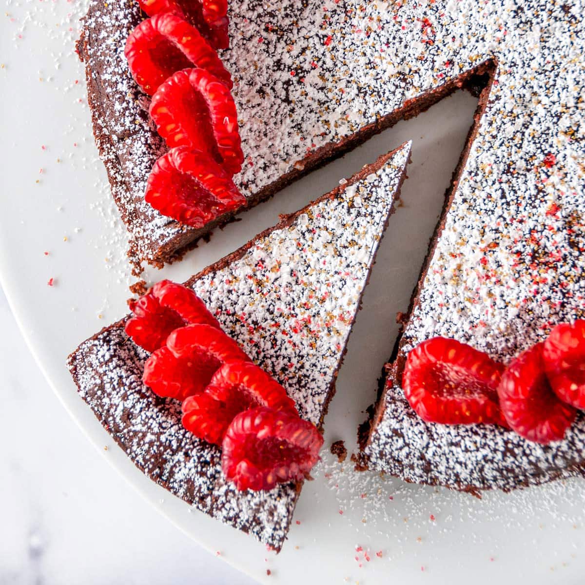 Chocolate Raspberry Flourless Cake sprinkled with powdered sugar on white cake stand with one slice cut overhead view