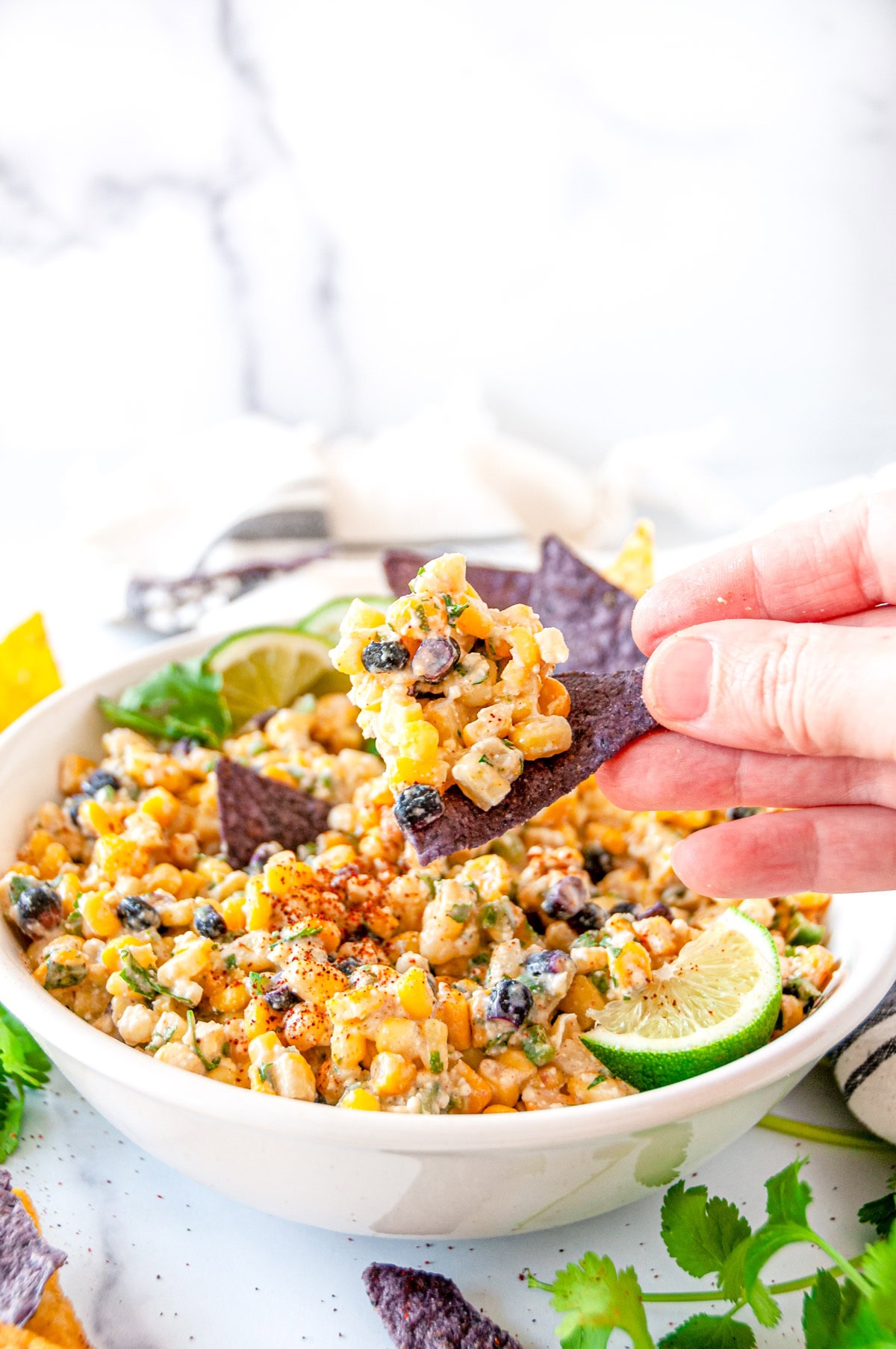 Elote dip in white bowl with purple and yellow tortilla chips with a bite held in hands side view close up
