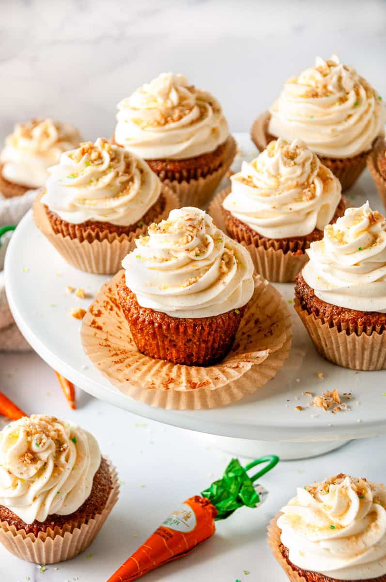 Carrot Cake Cupcakes with cream cheese frosting on a white cake stand, topped with chopped nuts and sprinkles