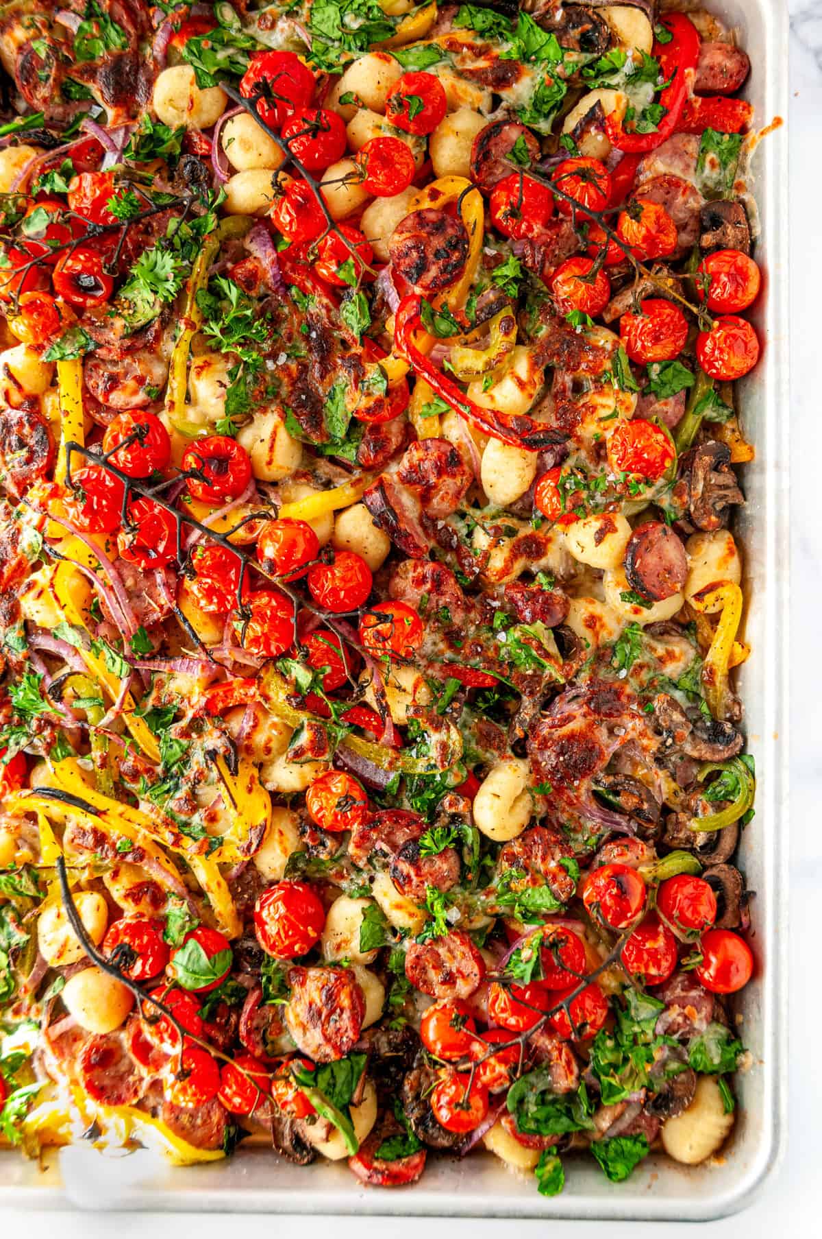 Overhead view of finished sheet pan sausage gnocchi with roasted vegetables, melted cheese, and fresh herbs.