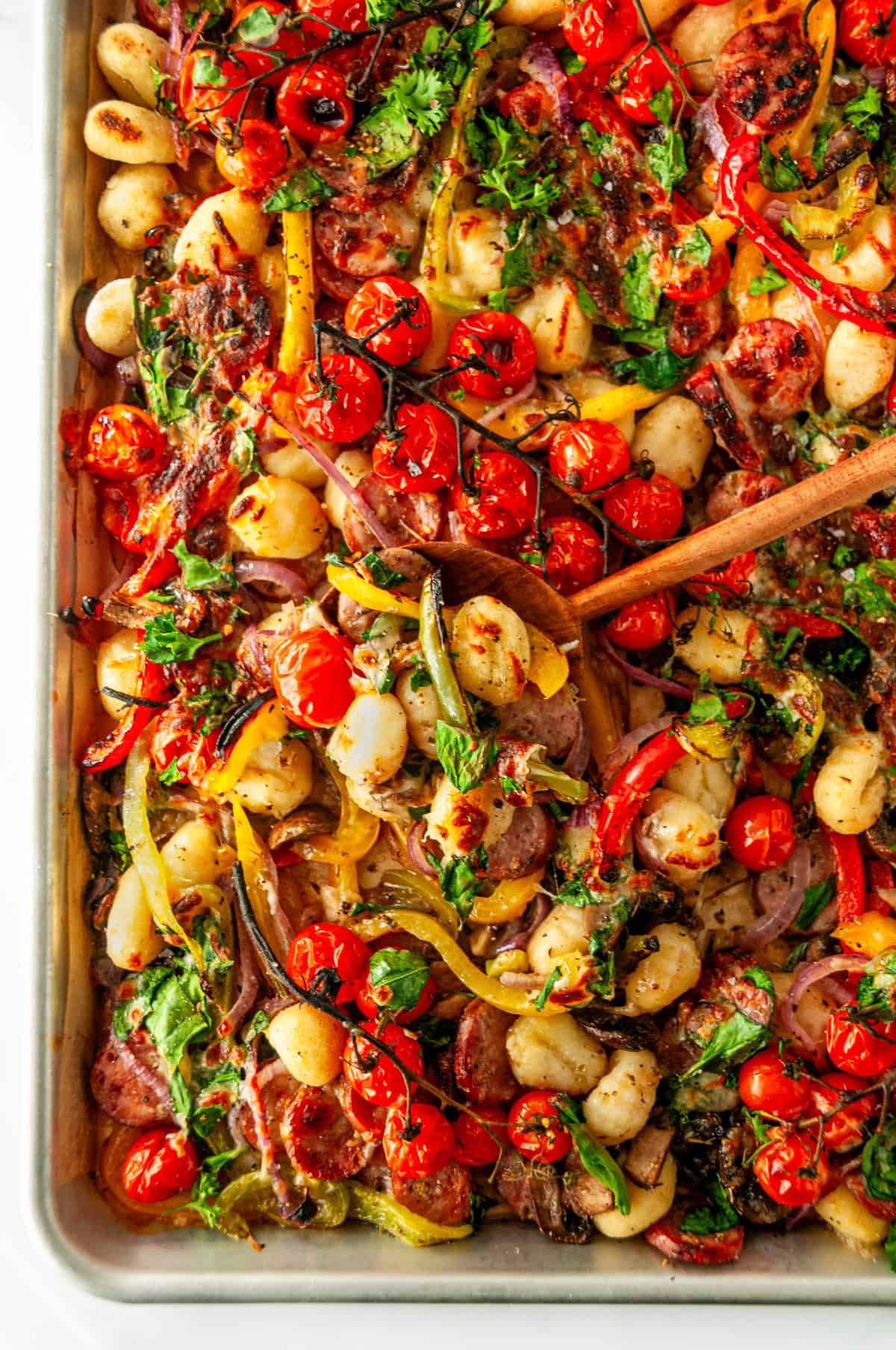 Vertical view of finished sheet pan sausage gnocchi with roasted peppers, tomatoes, gnocchi, and melted cheese with wooden spoon.