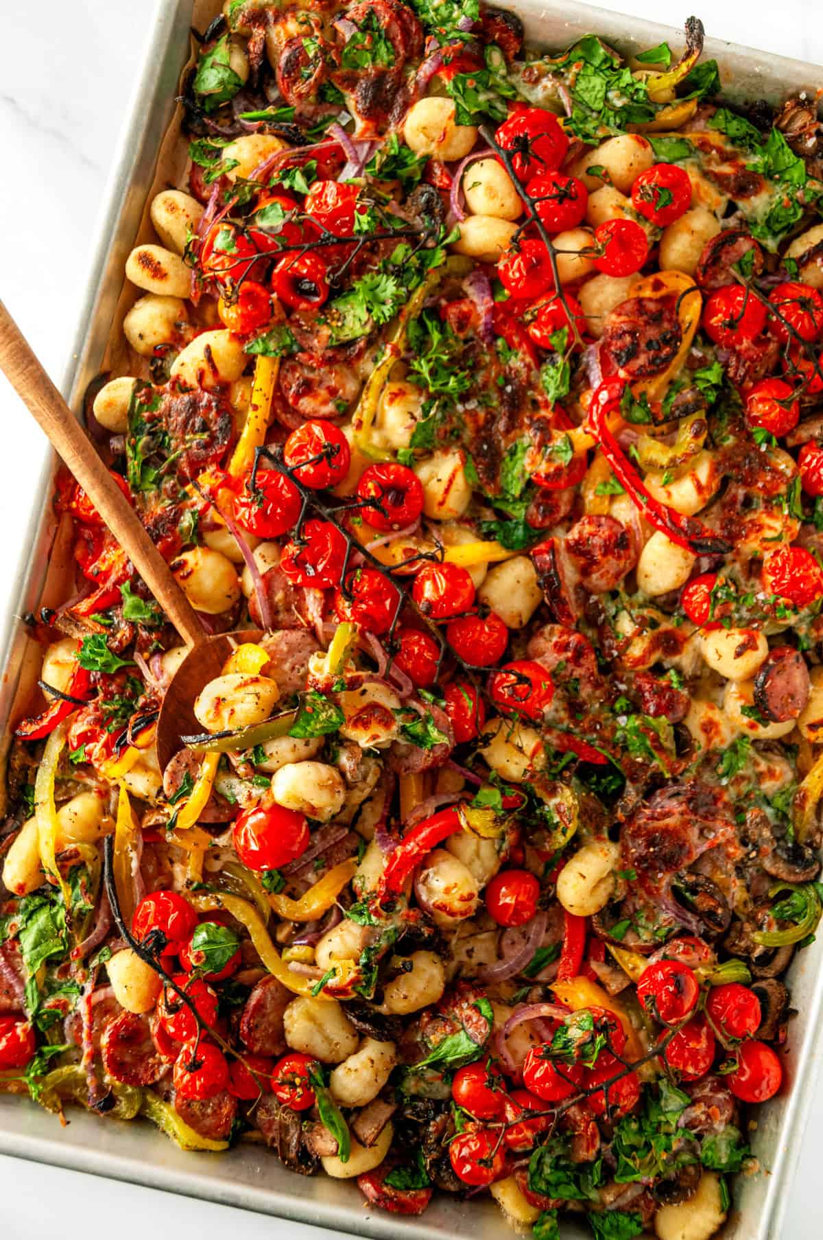 Sheet pan sausage gnocchi with a wooden spoon lifting a serving of gnocchi, turkey sausage, roasted vegetables, and melted cheese.