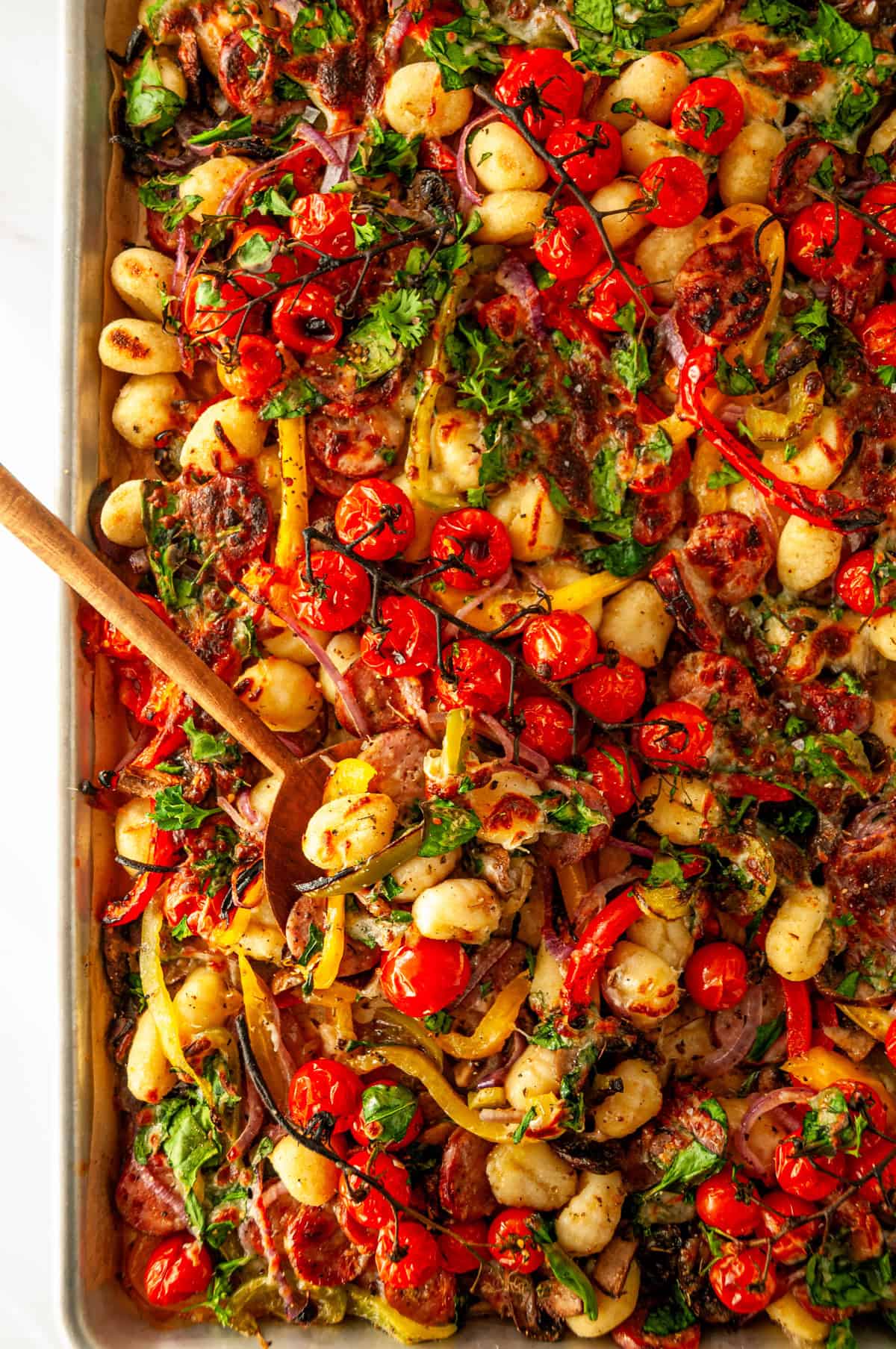 Overhead angle of sheet pan sausage gnocchi with a wooden spoon and roasted tomatoes, peppers, gnocchi, and turkey sausage.
