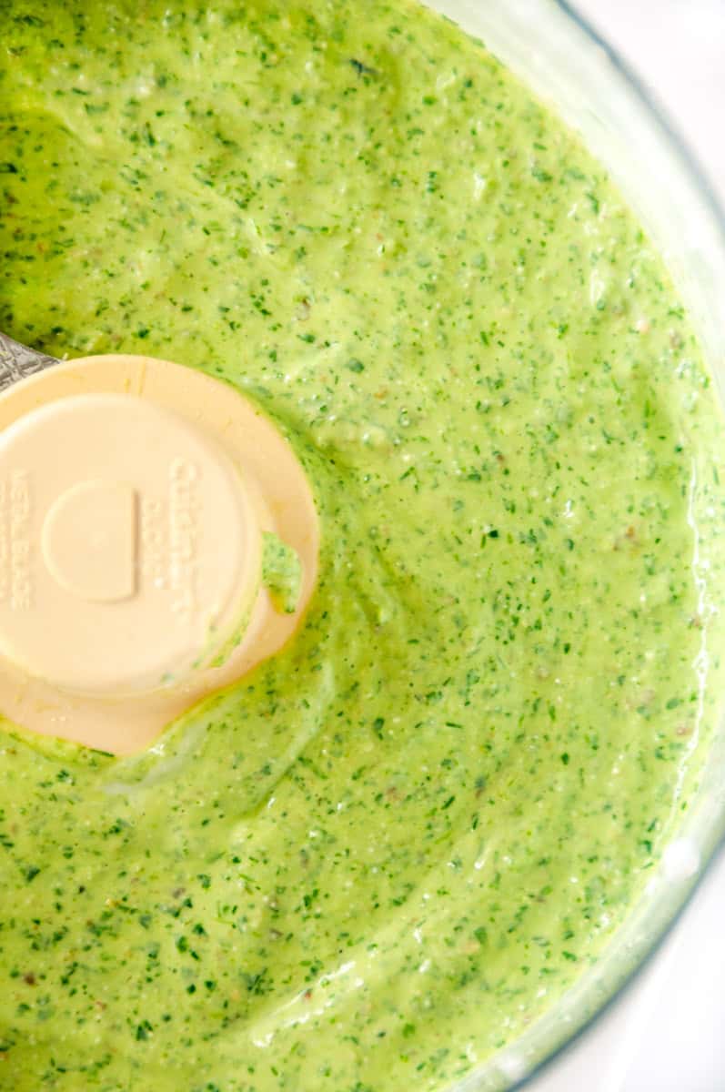 Close-up of creamy green goddess dressing in a food processor with flecks of fresh herbs.