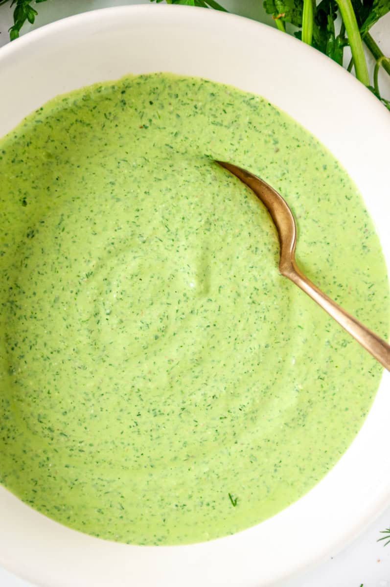 Green goddess dressing in a white bowl with a gold spoon, surrounded by fresh herbs.