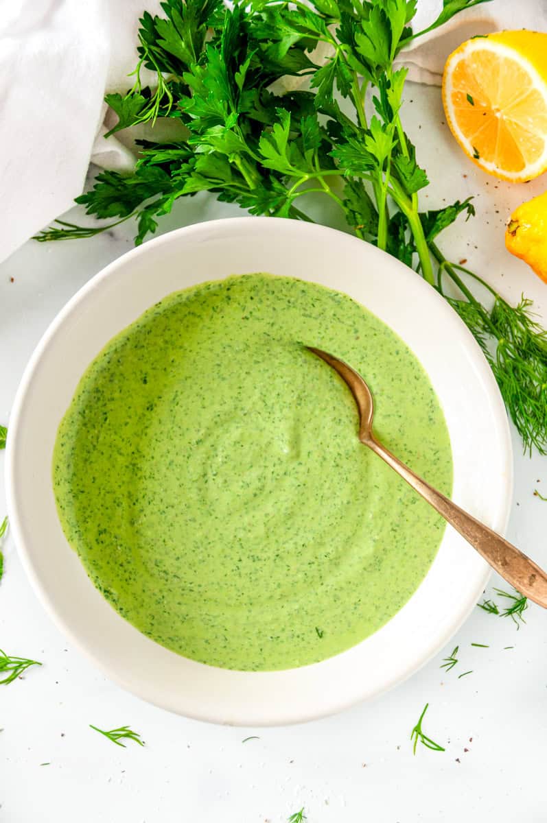 Bowl of creamy green goddess dressing with fresh herbs and lemon on a white surface
