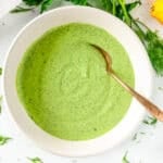 Bowl of creamy green goddess dressing with fresh herbs and lemon on a white surface
