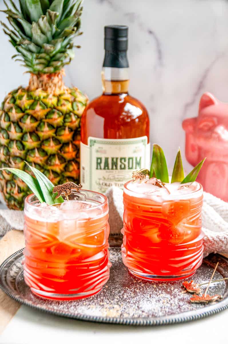 Jungle Cat cocktail with a whole pineapple, a bottle of Ransom, and a pink ceramic cat on a white marble counter.