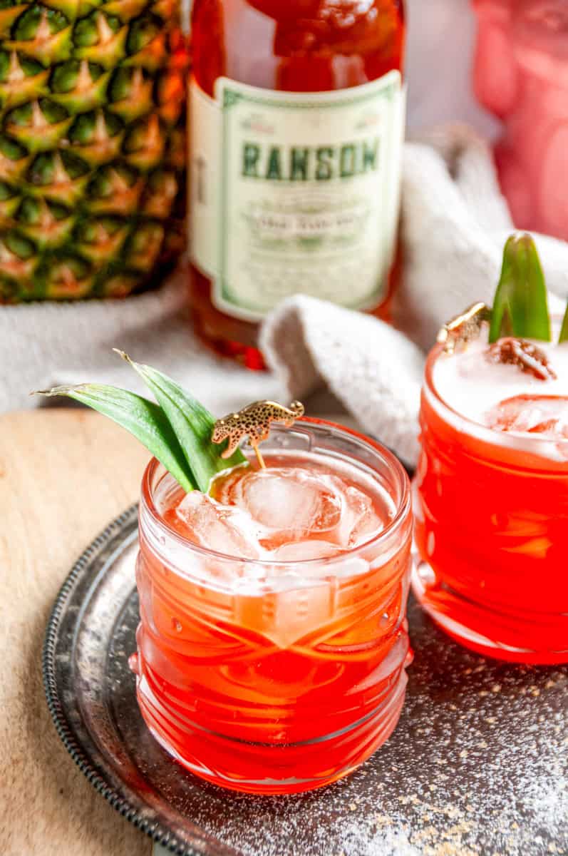 Close-up of a Jungle Cat cocktail garnished with pineapple leaves, with a bottle of Ransom and a fresh pineapple behind it.