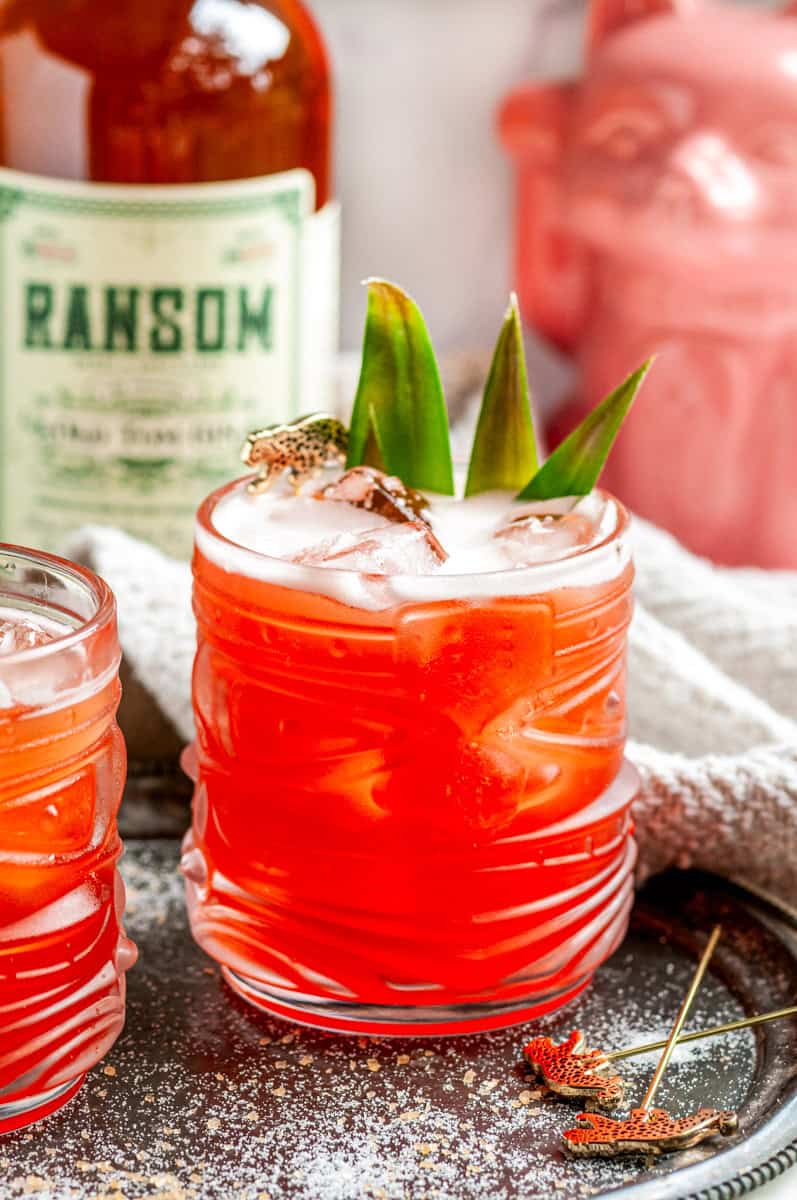 Bright red Jungle Cat cocktail over ice, garnished with pineapple leaves and a leopard pick, next to a pink ceramic cat.