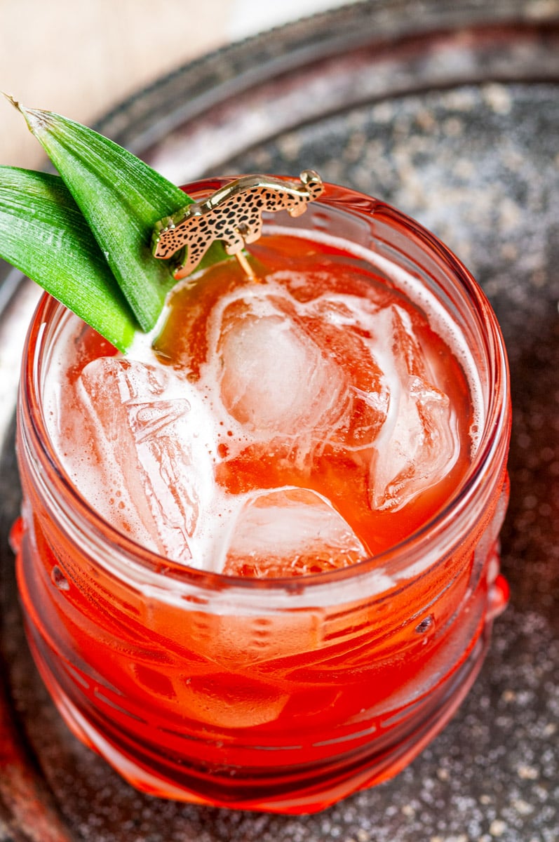 Overhead view of a red Jungle Cat cocktail over ice with pineapple leaves and a leopard garnish on a metal tray.